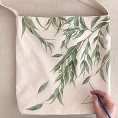 a person is painting a bag with green leaves on the front and back side, while holding a pencil in their left hand