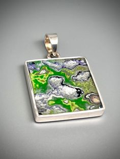 a square silver pendant with green and blue designs on the front, sitting on a gray surface