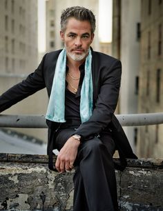 a man in a black suit and blue scarf sitting on a ledge looking at the camera