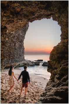 two people walking on the beach holding hands and looking at the ocean through a cave