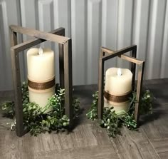 two white candles sitting next to each other on top of a wooden table with greenery