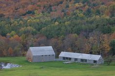 two barns sit in the middle of a green field with fall trees behind them and a pond