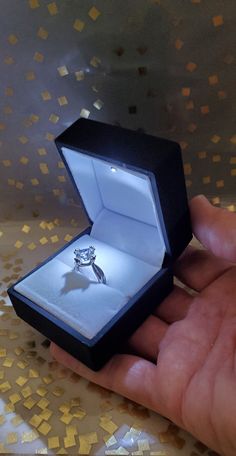 a hand holding an open ring box with a white diamond in it's center