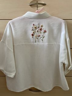 a white shirt with flowers embroidered on the front and back, hanging from a wooden hanger