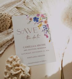 a wedding save the date card with flowers and feathers next to some seashells