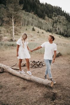 a man and woman holding hands while walking across a log in the dirt near a forest