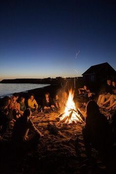 a group of people sitting around a campfire on the beach at night with one person standing