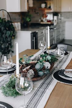 the table is set for christmas dinner with white candles and greenery on each side