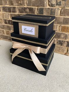 three black boxes with gold trim and ribbons are stacked on top of each other in front of a brick wall