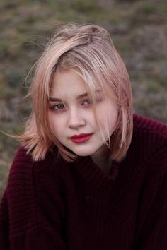 a woman with blonde hair and blue eyes wearing a red sweater sitting on the ground