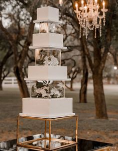 a tall white cake with flowers on top sitting in front of a chandelier