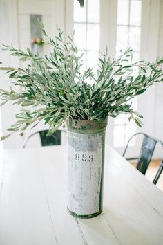 a white table topped with a metal can filled with green plants and greenery next to a window