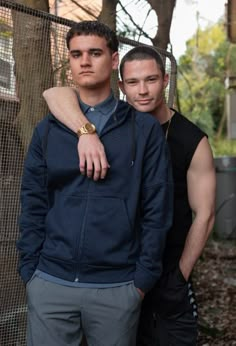 two young men standing next to each other in front of a chain link fence and trees