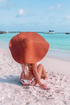 a woman sitting on top of a sandy beach next to the ocean wearing a large orange hat