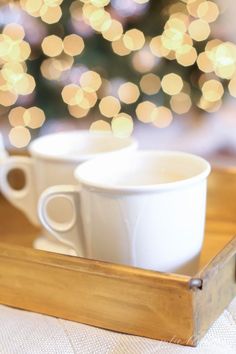 two white coffee cups sitting on top of a wooden tray next to a christmas tree