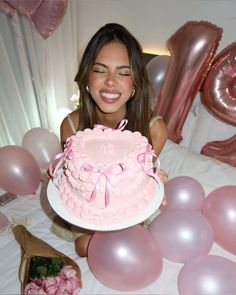 a woman holding a pink birthday cake in front of balloons