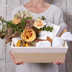 a woman holding a wooden box with flowers and other items in it while wearing a gray sweater