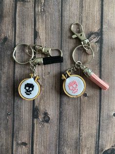 two cross stitch keychains are on a wooden surface, one has a skull and the other has a pencil