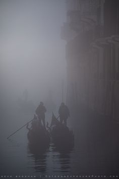 two people on boats in the foggy water with buildings and balconies behind them