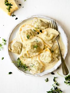 a white plate topped with ravioli next to a slice of bread and green herbs