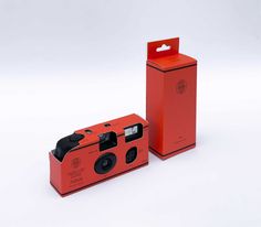 an orange camera sitting in front of a red box on a white surface with its lid open