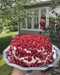 someone holding up a cake with strawberries on it in front of a house and flowers