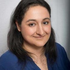 a woman with long black hair wearing a blue shirt and smiling at the camera while standing in front of a white wall