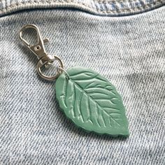 a green leaf shaped keychain hanging from a pair of jeans with a metal hook