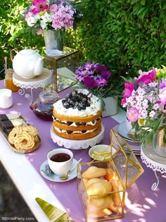 a table topped with cakes and flowers on top of a purple table cloth covered in food