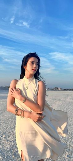 a woman standing in the sand wearing a white dress and gold bracelets with her arms crossed