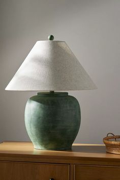 a green vase sitting on top of a wooden table next to a white shade lamp