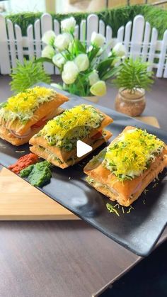 three small sandwiches on a black plate with flowers in the backgroung area