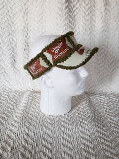 a white mannequin head wearing a green crocheted visor with two coca - cola cans on it