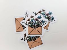 three envelope stickers with flowers in them on a white surface, one is brown and the other is blue