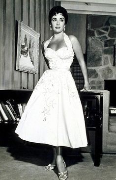 a woman in a white dress standing next to a book shelf with books on it