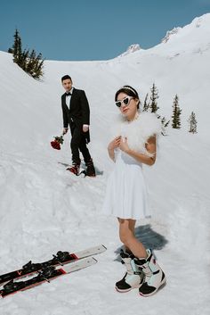 a man and woman standing on top of a snow covered slope next to skis