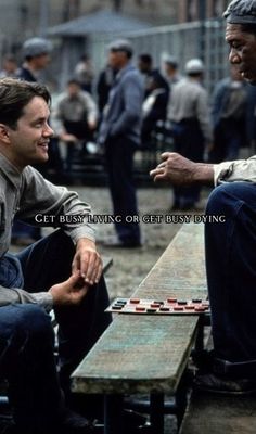 two men sitting at a picnic table playing checkers with one man in the background