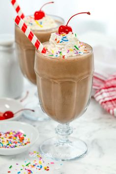 two glasses filled with chocolate milkshake and topped with whipped cream, sprinkles and cherries