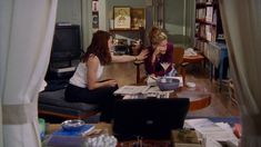 two women sitting at a table in a living room with laptops and books on it