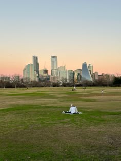 a man sitting on top of a lush green field next to a tall city skyline