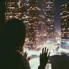 a woman standing in front of a window looking at the city lights and skyscrapers