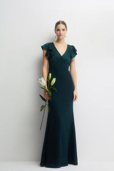 a woman in a long green dress holding a flower and standing next to a white wall