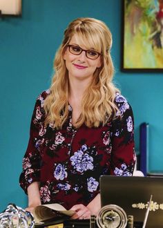 a woman with glasses standing in front of a laptop computer and looking at the camera