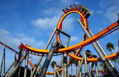 an orange roller coaster in the middle of a blue sky with palm trees behind it