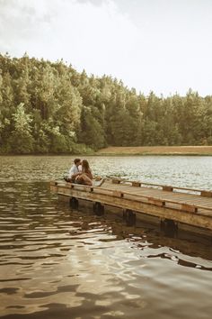 two people are sitting on a dock in the middle of a body of water with trees in the background