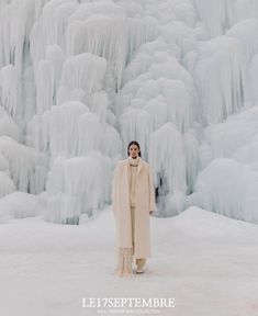 a woman standing in front of an ice - covered waterfall wearing a long beige coat