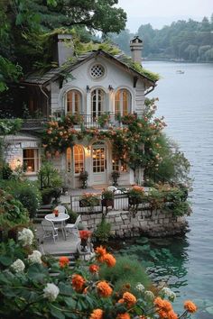 the house is surrounded by greenery and flowers on the water's edge, with boats in the background