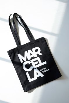 A canvas bucket tote bag in 100% European cotton, with a bottom panel for added capacity. The tote features an oversized MARCELLA NEW YORK graphic on one side, in white print on black canvas.[SPLIT] Our production team enjoys a living wage, a 40-hour work week with paid overtime, free health insurance and 20 days annual paid vacation. Black Shoulder Bag With Logo Print For Travel, White Shoulder Bag With Logo For Everyday Use, Black Shoulder Bag With Logo Print For Everyday, Black Shoulder Bag With Logo Print, White Shoulder Bag With Logo Print, Casual Shoulder Bag With Logo Print For Travel, Casual Travel Shoulder Bag With Logo Print, Casual Logo Print Shoulder Bag For Travel, Canvas Tote Shoulder Bag With Logo Print