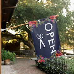 an open sign hanging from the side of a building with flowers on it and a pair of scissors