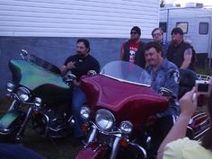 a group of men sitting on motorcycles in front of a house with one man taking a photo